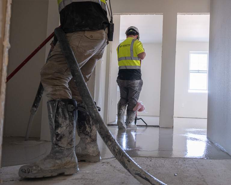 Construction workers in safety gear and boots pouring and smoothing a gypsum underlayment floor in a Minnesota multifamily residential building.