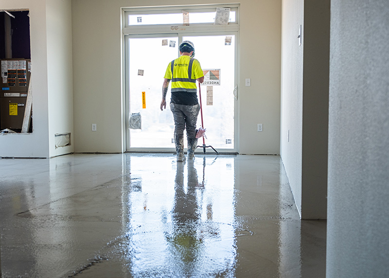 A construction worker in a high-visibility vest using a placer rake to smooth and distribute fresh Gyp-Crete 2000 gypsum underlayment over an apartment floor in Minnesota.