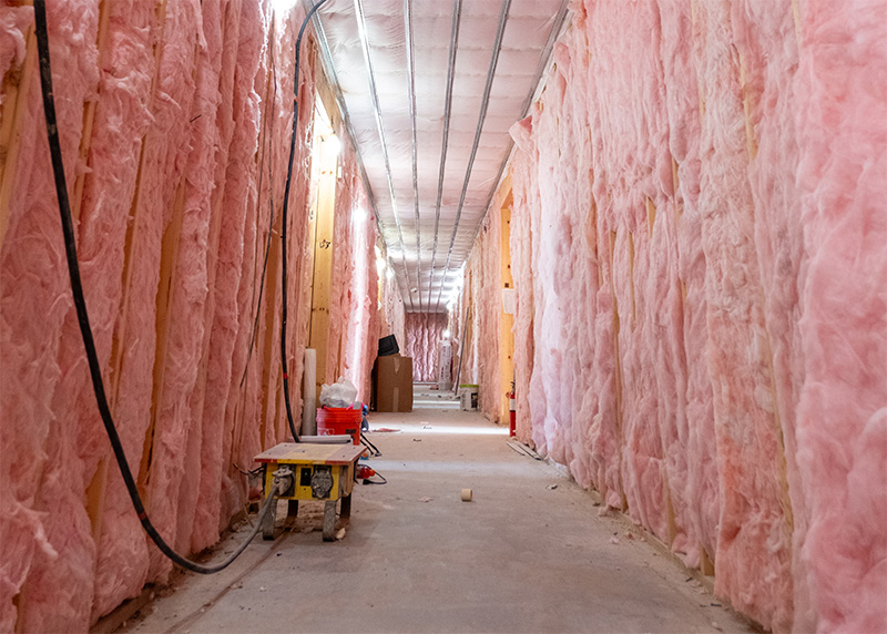 A long hallway inside a multifamily apartment complex under construction with pink fiberglass batt insulation completely filling the framed wall cavities in Minnesota.